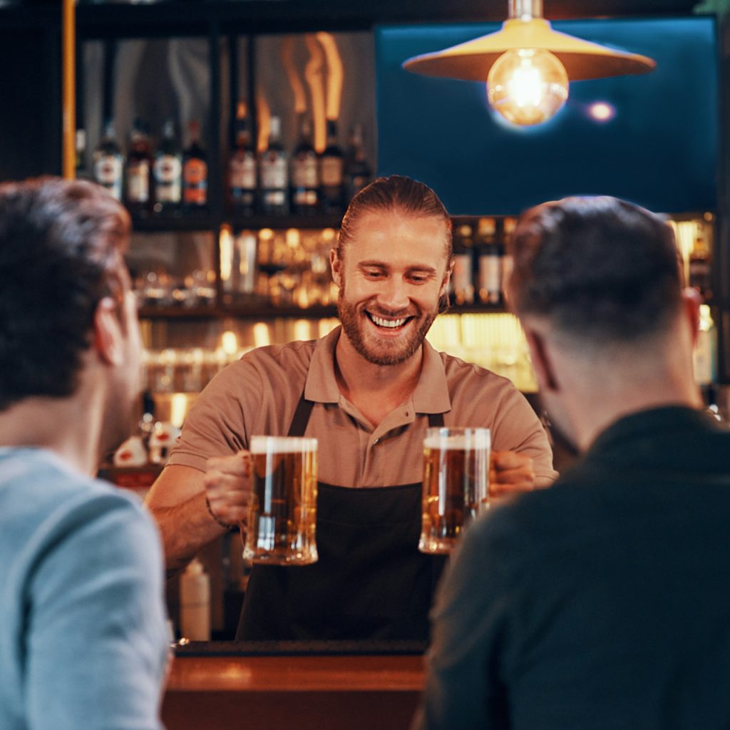 Cheerful bartender serving beer to young men while standing at the bar counter in pub